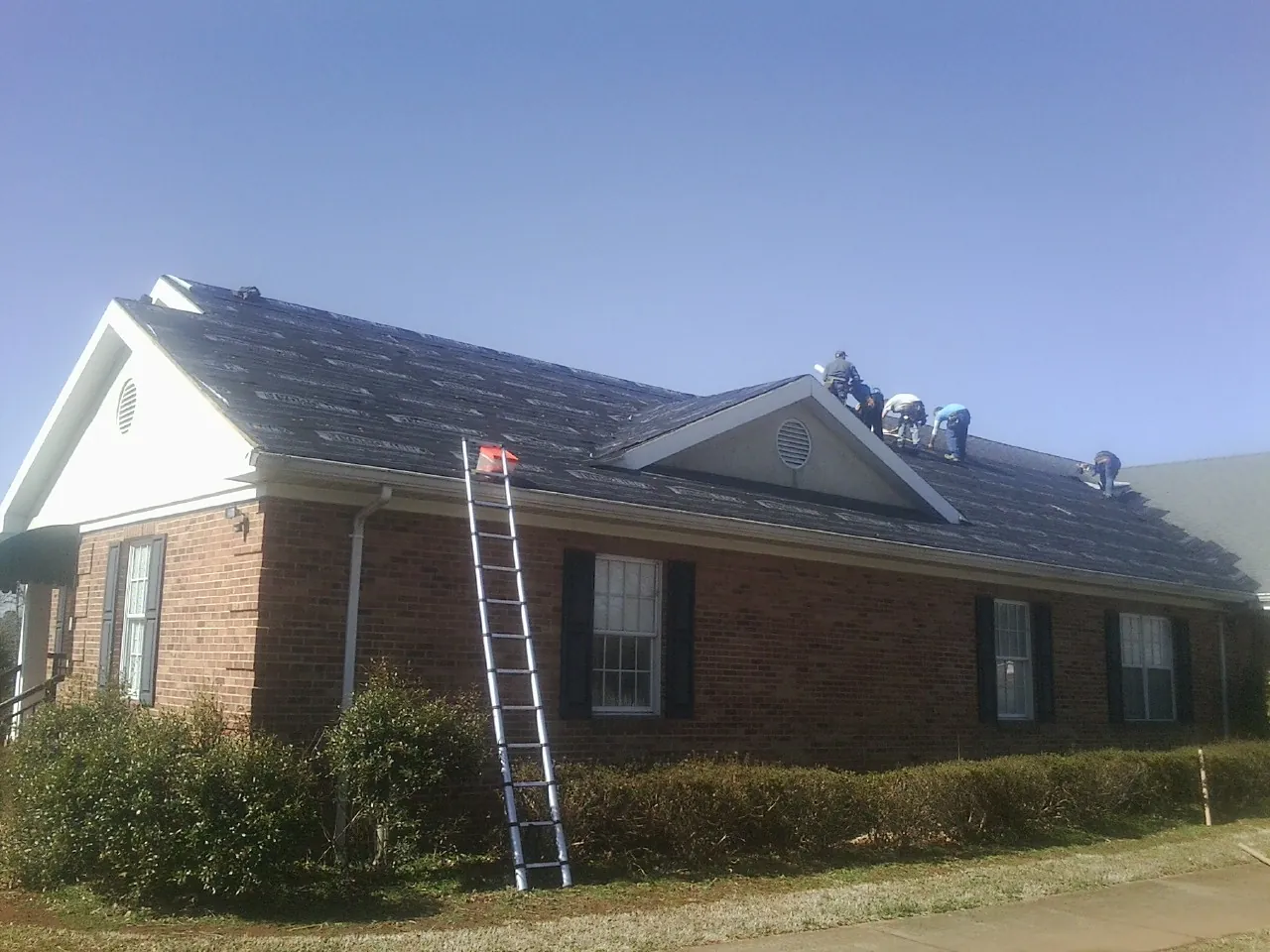 Crew tearing shingles off a brick ranch roof with a ladder set — Emergency Roof Repair work by our crew in Newalla