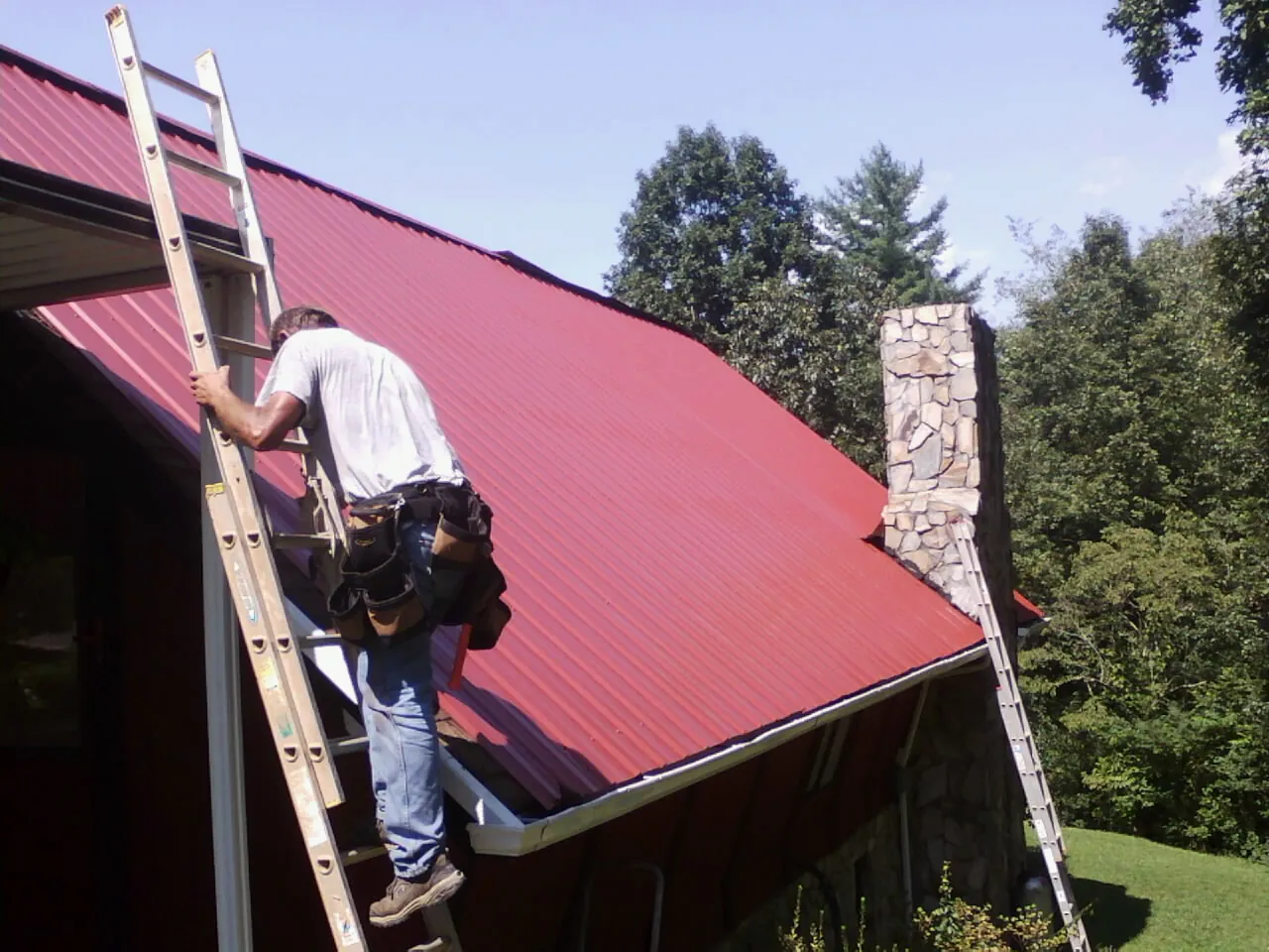 Roofer on a ladder beside a red metal roof and stone chimney — Soffit & Fascia Repair by Roofing Brothers in Newalla